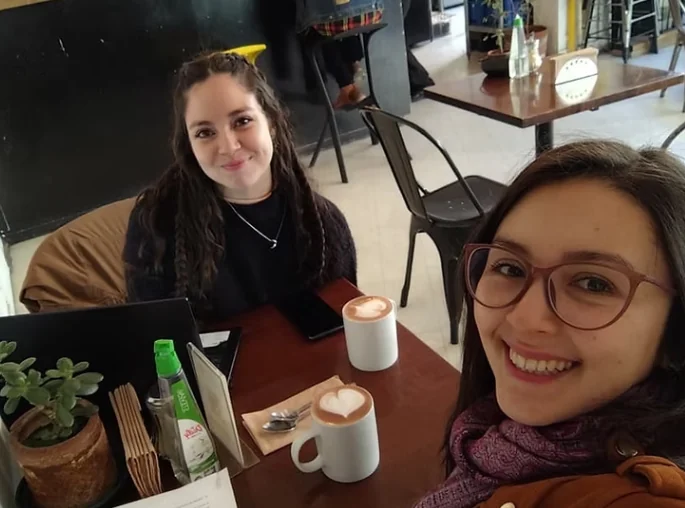 Yaz y Cami sonríen en una cafetería, con dos cafés con arte latte sobre la mesa.